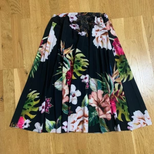 Blommig plisserad kjol från New Style - Säljer en svart plisserad kjol med stort blommigt mönster i grönt, rosa, vitt och rött från New Style Italy. Kjolen har elastisk midja och är perfekt för dig som gillar färgstarka prints och flowig känsla.