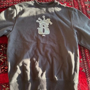 Svart sweatshirt från Stüssy med krona - Säljer en svart sweatshirt från Stüssy med stort S och krona tryckt på bröstet. Tröjan har rund halsringning och ribbade muddar vid ärmslut och nederkant. Perfekt för en avslappnad stil. Rare tryck, Fint skick