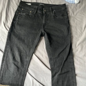 Svarta jeans från jack and jones stl 30/32 - Säljer ett par svarta jeansbyxor från Jack and Jones med klassisk femficksdesign och raka ben. Byxorna har knappgylf och diskreta sömmar, perfekta för en stilren look. Byxorna är skinny 