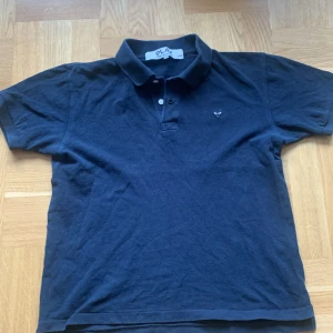 Svart pikétröja från Comme des Garçons Play - Säljer en svart pikétröja från Comme des Garçons Play med klassisk krage och två knappar framtill. Tröjan har det ikoniska hjärte-logotypen broderad på bröstet. Perfekt för en stilren och avslappnad look.