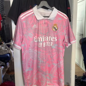 Real Madrid rosa fotbollströja med drakmönster - Säljer en rosa Real Madrid fotbollströja från Adidas med vitt drakmönster och vit krage. Tröjan har korta ärmar, klubbmärke på bröstet och Adidas-logga. Materialet är lätt och andas, perfekt för träning eller match. Använd en gång och har ett litet märke. Köpen fel storlek