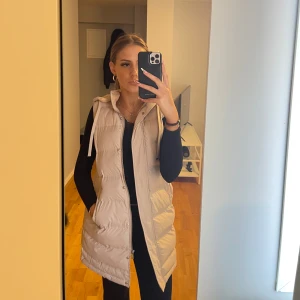 Beige lång väst med luva - Säljer en beige lång dunväst från Fransa med huva och dragsko. Västen har två fickor framtill och stängs med dragkedja eller knappar. Perfekt till vår/sommar och att slänga över en hoodie eller tröja för extra värme.