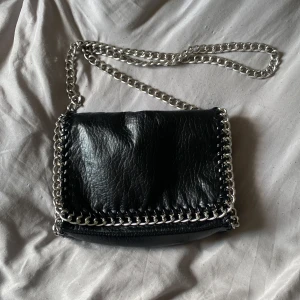Svart axelväska med kedjedetaljer från Tiamo - Säljer en svart axelväska från Tiamo i skinnimitation med silverfärgad kedja som axelrem och runt kanterna. Väskan har magnetknappar och ett innerfack med dragkedja.  28x19