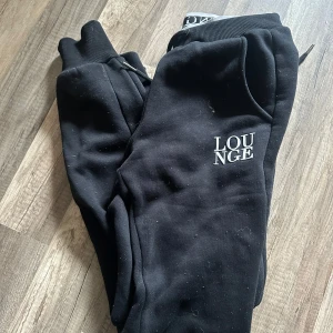 Svarta mjukisbyxor från Lounge - Svarta mjukisbyxor från Lounge med vit logga på ena benet. Byxorna har resår i midjan och vid bensluten samt dragsko för justerbar passform. Perfekta för en avslappnad stil. Noppiga men går nog få bort 