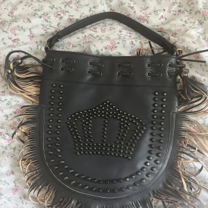 Svart axelväska med nitar och fransar - Cool svart axelväska i skinnimitation med stora nitar som bildar en krona på framsidan. Väskan har långa fransar i svart och beige längs kanterna och snörningsdetaljer upptill. Får plats med mycket och inte använd, skriv för fler bilder mm