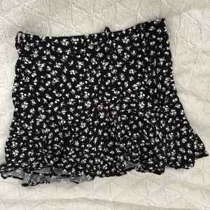 Svart blommig kjol från Zara - Supersöt svart kjol från Zara med små vita blommor och volang nertill. Kjolen har en kort längd och en lätt utsvängd form som ger en luftig känsla. Perfekt att styla med en enkel topp eller hoodie. Kjolen har även inbyggda shorts som som ger den en extra säker känsla. Kjolen säljs då den är lite stor för mig som är XS och kommer då tyvärr inte till användning.