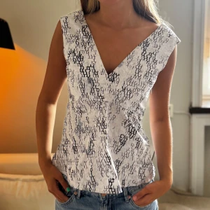 Vit mönstrad omlottopp - Säljer en vit omlottopp med svart abstrakt mönster. Toppen har djup v-ringning både fram och bak och är ärmlös. Perfekt till jeans för en snygg och modern look.