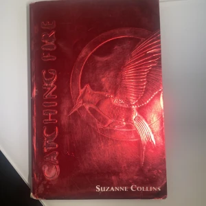 Catching Fire - Säljer boken 'Catching Fire' av Suzanne Collins. Omslaget är metallicrött med en präglad symbol av en fågel och text i silver. Boken är på engelska och tillhör Hunger Games-serien. Boken är på engelska! 🤍