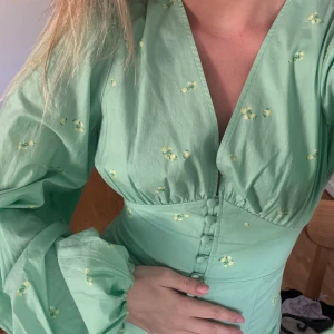 Blommig adoore-klänning - Superfin mintgrön klänning med små gula blommor, djup v-ringning och markerad midja. Klänningen har långa puffärmar och knappar framtill. Perfekt för vår och sommar! Storlek 34.