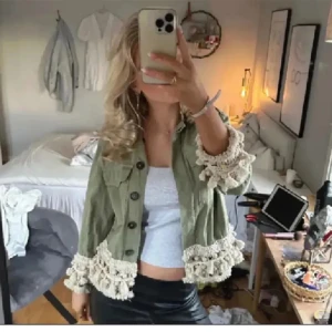 Grön jacka med volang och pärldetaljer - Säljer en ljusgrön jacka med stora knappar och dekorativa volanger samt pärlor och spets längs ärmslut och nederkant. Jackan har två bröstfickor och är croppad i modellen. Perfekt för dig som vill sticka ut lite extra!