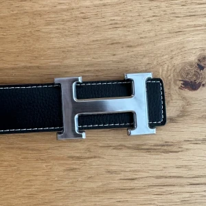 Svart skärp med H-spänne - Säljer ett svart skärp med ett stort, silverfärgat H-spänne. Skärpet har vit kontrastsöm längs kanterna och är gjort i skinn. Perfekt accessoar för att lyfta din outfit.