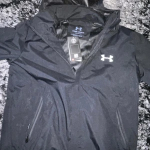 Svart vindjacka från Under Armour - Säljer en svart vindjacka från Under Armour med en ficka på bröstet och två sidofickor. Jackan har en sportig look med logga på bröstet och texten 'Explore' på ena sidan. Perfekt för utomhusaktiviteter och träning. (Helt nytt med kartong på)