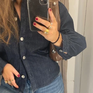 Mörkblå jeansskjorta med silverknappar - Snygg mörkblå jeansskjorta med stora silvriga knappar framtill. Skjortan har lång ärm och klassisk krage, perfekt till jeans eller kjol för en cool look. Materialet är denim och passformen ser normal ut.
