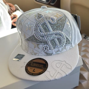 Vit keps med glittriga detaljer och dollartecken - Helt ny vit keps från Leader med stora glittriga silverfärgade dollar- och stjärndetaljer framtill. Broderat dollartecken även baktill. Kepsen har platt skärm, den är som 59Fiffty modellerna och har 56-57 i storlek ungäfer. 