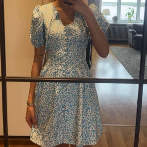 Blommig blå kortklänning från Trendyol - Supersöt blå klänning med vita blommor från Trendyol. Klänningen har korta puffärmar, v-ringning och knappar framtill. Den har en figurnära passform upptill och utställd kjol. Perfekt för vår och sommar!