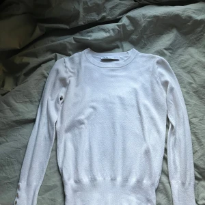Vit stickad tröja med knappar från Zara - Säljer en vit stickad tröja från Zara med rund halsringning och långa ärmar. Tröjan har dekorativa knappar i guld på båda ärmarna och en smal, figurnära passform. Perfekt för sena och kalla sommarkvällar 