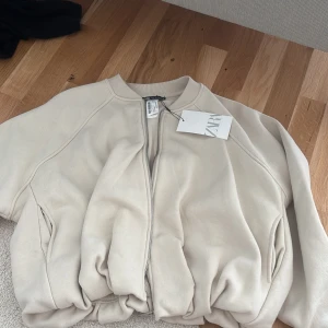 Beige bomberjacka från Zara - Säljer en beige bomberjacka från Zara med dragkedja framtill. Jackan har en oversized passform och ribbade muddar vid hals och ärmslut. Perfekt för att lyfta din outfit med en trendig touch.