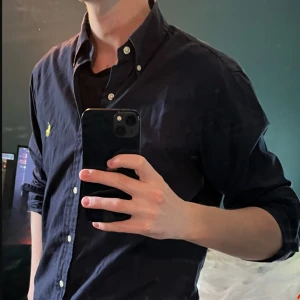 Mörkblå skjorta från Ralph Lauren - Säljer en klassisk mörkblå skjorta från Ralph Lauren med knappar framtill och gul broderad logga på bröstet. Skjortan är i storlek S och är i perfekt passform för 170 - 180