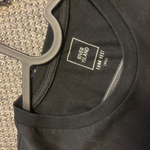 Svart linnetopp från River Island - Säljer en svart ärmlös linnetopp från River Island i storlek S. Toppen har rund halsringning och är enkel i modellen, perfekt för en stilren look.