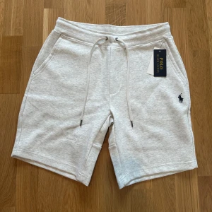 Grå shorts från Polo Ralph Lauren - Snygga ljusgrå shorts från Polo Ralph Lauren med dragsko i midjan och klassisk logga broderad på ena benet. Mjukt bomullsmaterial, två sidofickor och en bakficka. Perfekta för chill dagar eller när du vill ha en clean look.