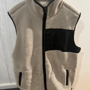 Beige fleeceväst från Newbody - Mysig beige fleeceväst från Newbody med svarta detaljer och dragkedja framtill. Västen har hög krage, två fickor i sidorna och en bröstficka med dragkedja. Perfekt lager-på-lager-plagg för kyliga dagar.