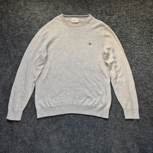 Beige stickad tröja från GANT - Snygg beige stickad tröja från GANT med broderad logga på bröstet. Tröjan har rund halsringning med diskret randig detalj på insidan. Perfekt för dig som gillar en clean och klassisk stil.