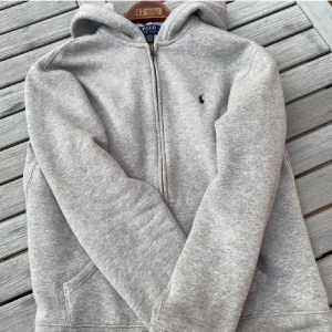 Grå hoodie från Polo Ralph Lauren XL - Klassisk grå hoodie från Polo Ralph Lauren i storlek XL. Tröjan har dragkedja, huva och känguruficka framtill. Liten broderad logga på bröstet. Perfekt för chill dagar och enkel att matcha med jeans eller joggers.