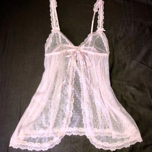 Rosa spets linne - Supersöt ljusrosa linnetopp i transparent mesh med små prickar och spetsdetaljer längs kanterna. Smala (justerbara) axelband, rosett framtill och v-ringning. Perfekt för lager-på-lager eller som statement piece. Toppens material är lätt och luftig syntet. 