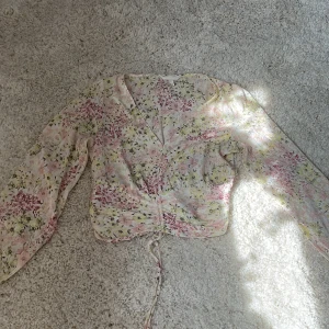 Blommig blus med snörning från H&M - Supersöt blommig blus från H&M i tunnt, luftigt material. Ljus botten med rosa, gula och gröna blommor, v-ringning och långa ärmar. Smockad baktill och snörning framtill för extra snygg passform. Perfekt för vår och sommar!