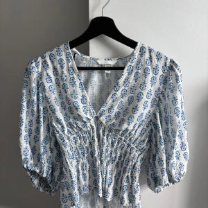 Vit och blå mönstrad klänning - Supersöt vit klänning med blått blommönster och v-ringning. Klänningen har puffiga trekvartsärmar och smockad midja som ger en snygg siluett. Perfekt för sommarens alla tillfällen! Använd en gång nyskick