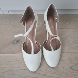 Vita pumps med klack från Anna Field - Stilrena vita pumps från Anna Field med rund tå och smal klack. Skorna har en elegant rem över foten med spänne och är tillverkade i syntetmaterial. Perfekta för dig som vill ha en klassisk och feminin look med en modern twist. Använda 1 gång på bröllop 