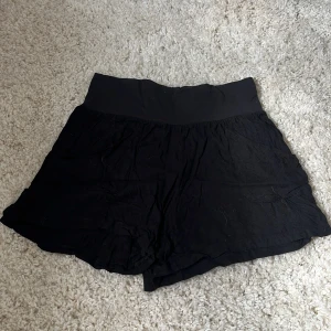 Svarta högmidjade shorts i bomull - Svarta, lösa shorts med bred resår i midjan för extra komfort. De är tillverkade i ett tunt och mjukt bomullstyg som känns luftigt och perfekt för varma dagar. Shortsen har en enkel och stilren design utan synliga detaljer eller mönster.