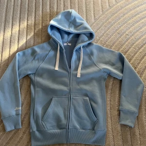 Ljusblå hoodie från Russemerch  - Ljusblå hoodie från Russemerch med dragkedja, vita snören i huvan och stora fickor framtill. Tröjan har ribbade muddar och är långärmad, perfekt för chill dagar. Materialet är mjuk bomullsmix som känns skönt mot huden.