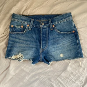 Levi's 501 jeansshorts med slitningar - Klassiska blå jeansshorts från Levi's modell 501 med råa, fransiga kanter och slitningar framtill. Shortsen har fem fickor, knappgylf och den ikoniska läderpatchen bak. Perfekta för en avslappnad och cool stil under sommaren.