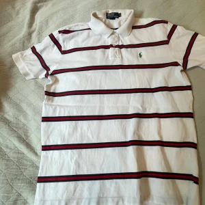 Randig piké från Polo Ralph Lauren - Vit pikétröja från Polo Ralph Lauren med klassisk krage och knappar. Tröjan har horisontella ränder i svart och rött samt den ikoniska gröna loggan broderad på bröstet. Perfekt för en stilren och sportig look.