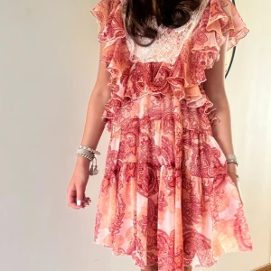 Röd paisleyklänning  - Superfin kortklänning från ASOS Design med röd paisleyprint och volanger. Klänningen har spetsdetaljer upptill, knytning i nacken och är tillverkad i ett lätt material. Perfekt för sommarens alla tillfällen 