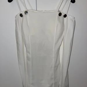 Vit långärmad klänning med axelband - Säljer en stilren vit klänning med långa ärmar och dubbla smala axelband. Klänningen har en rak passform och dekorativa knappar vid axlarna. Perfekt för dig som gillar minimalistisk och modern stil.