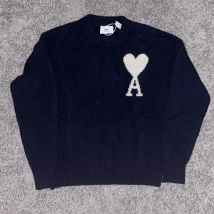 Mörkblå stickad tröja från Ami - Säljer en mörkblå stickad tröja från Ami Alexandre Mattiussi med vit hjärt- och A-detalj på bröstet. Tröjan har rund halsringning, ribbade muddar och är perfekt för en stilren look.