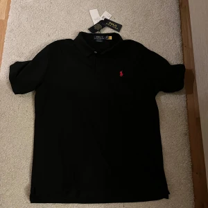 Svart pikétröja från Polo Ralph Lauren - Klassisk svart pikétröja från Polo Ralph Lauren med krage och knappar framtill. Tröjan har en liten röd broderad logga på bröstet och är tillverkad i bomull. Perfekt för en stilren och tidlös look.