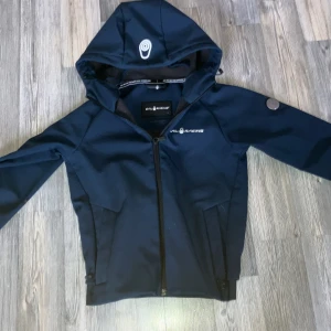 Marinblå softshelljacka från Sail Racing - Säljer en marinblå softshelljacka från Sail Racing med huva och hel dragkedja. Jackan har två sidofickor med dragkedja och en logotyp på bröstet. Perfekt för blåsiga dagar och snygg till vardags.