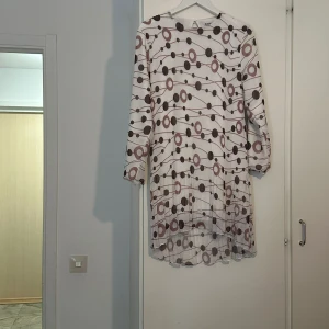 Vit och brun mönstrad långärmad klänning - Säljer en vit klänning med brunt och rosa prickigt och randigt mönster. Klänningen har lång ärm och är något längre i modellen. Perfekt för dig som gillar unika mönster och en avslappnad stil.