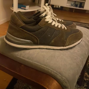 Bruna sneakers med mockadetaljer - Snygga bruna mocka  sneakers med paneler i textil, vita snören och vit sula med silverdetalj på hälen. Skorna har en klassisk rund tå och platt sula, perfekta för en avslappnad streetstil.