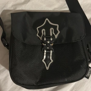 Svart axelväska från Trapstar - Snygg svart axelväska från Trapstar med justerbar rem och coolt metallspänne framtill. Väskan har en strukturerad yta och tydlig Trapstar-logga på både rem och etikett. Perfekt för att bära dina essentials med stil.