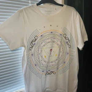 Vit t-shirt med grafiskt tryck från Guess - Vit t-shirt från Guess med stort grafiskt tryck på framsidan i flera färger, bland annat rött, gult och blått. Trycket har geometriska mönster, stjärnor och ormar. Klassisk rund hals och korta ärmar. Skön och lätt i bomull, perfekt för en cool och avslappnad stil.