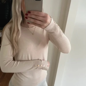 Beige långärmad tröja med knappar - Snygg beige långärmad tröja med smal passform och dekorativa guldfärgade knappar vid ärmsluten. Enkel och stilren design som passar perfekt till jeans eller kjol. Mjuk och skön känsla, perfekt för lager-på-lager.