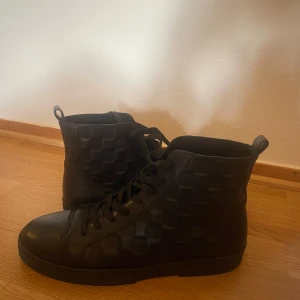 Svarta Louis Vuitton sneakers i skinn - Snygga svarta high-top sneakers från Louis Vuitton med schackrutigt mönster på sidorna. Skorna är tillverkade i skinn och har snörning framtill. Perfekt för dig som vill ha en stilren och exklusiv look med streetkänsla.