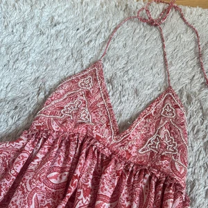 Rosa paisleymönstrad kortklänning - Supersöt kortklänning i rött och vitt med paisleymönster och tunna halterneckband. Klänningen har v-ringad omlott-topp med pärldetaljer och ett luftigt fall. Perfekt för varma dagar och sommarvibbar!