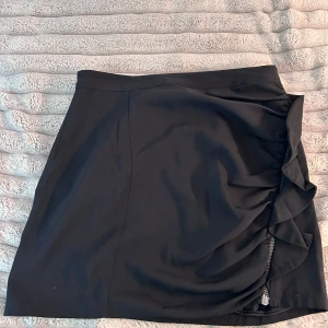 Svart kjol med dragkedja från Zara - Svart kortkjol från Zara Basic med snygg drapering och silverfärgad dragkedja längs sidan. Kjolen har en stilren look med en liten volangdetalj vid dragkedjan. Perfekt för dig som gillar en enkel men ändå cool stil.