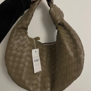 Olivgrön axelväska med flätad design  - Säljer en olivgrön axelväska / braided handbag / flätad handväska från Pitaya Teah Bag med snygg flätad design och rundad form. Väskan har en bred axelrem med knutdetalj och stängs med dragkedja. Perfekt för att lyfta din outfit med en trendig accessoar. Ny med prislapp kvar. 