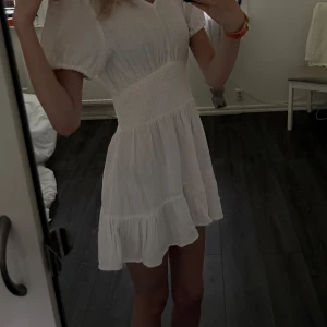 Vit, kortklänning med romantiska volanger ✨Aldrig använd med prislapp kvar. Passar bäst för dig som bär storlek XS. - Säljer en söt vit kortklänning med volangdetaljer och smock i midjan. Klänningen har korta puffärmar och v-ringad hals. Perfekt för sommarens varma dagar och har en luftig och lätt känsla.